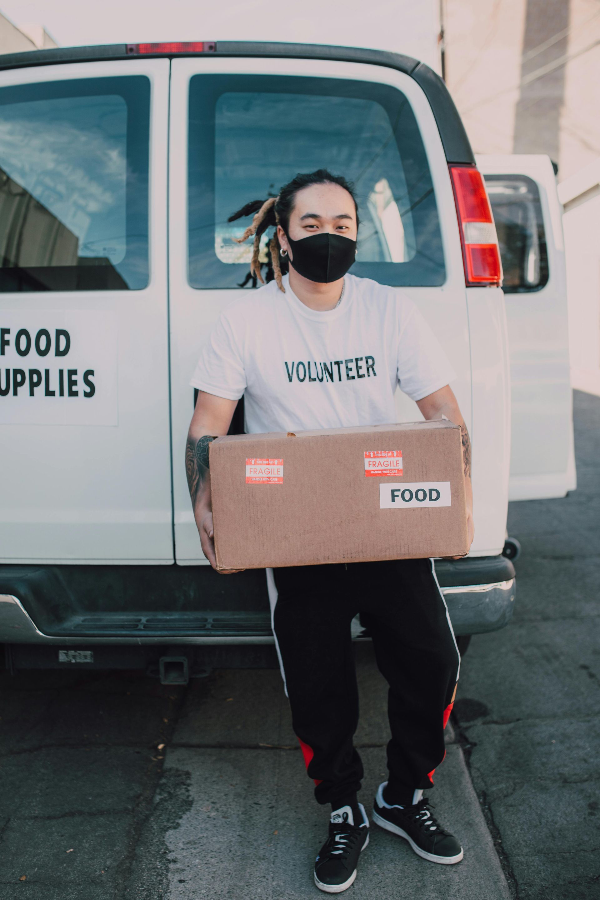 A man wearing a mask is holding a box in front of a van that says food supplies.