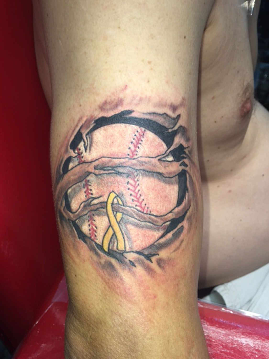 A man has a tattoo of a baseball with a yellow ribbon around it.