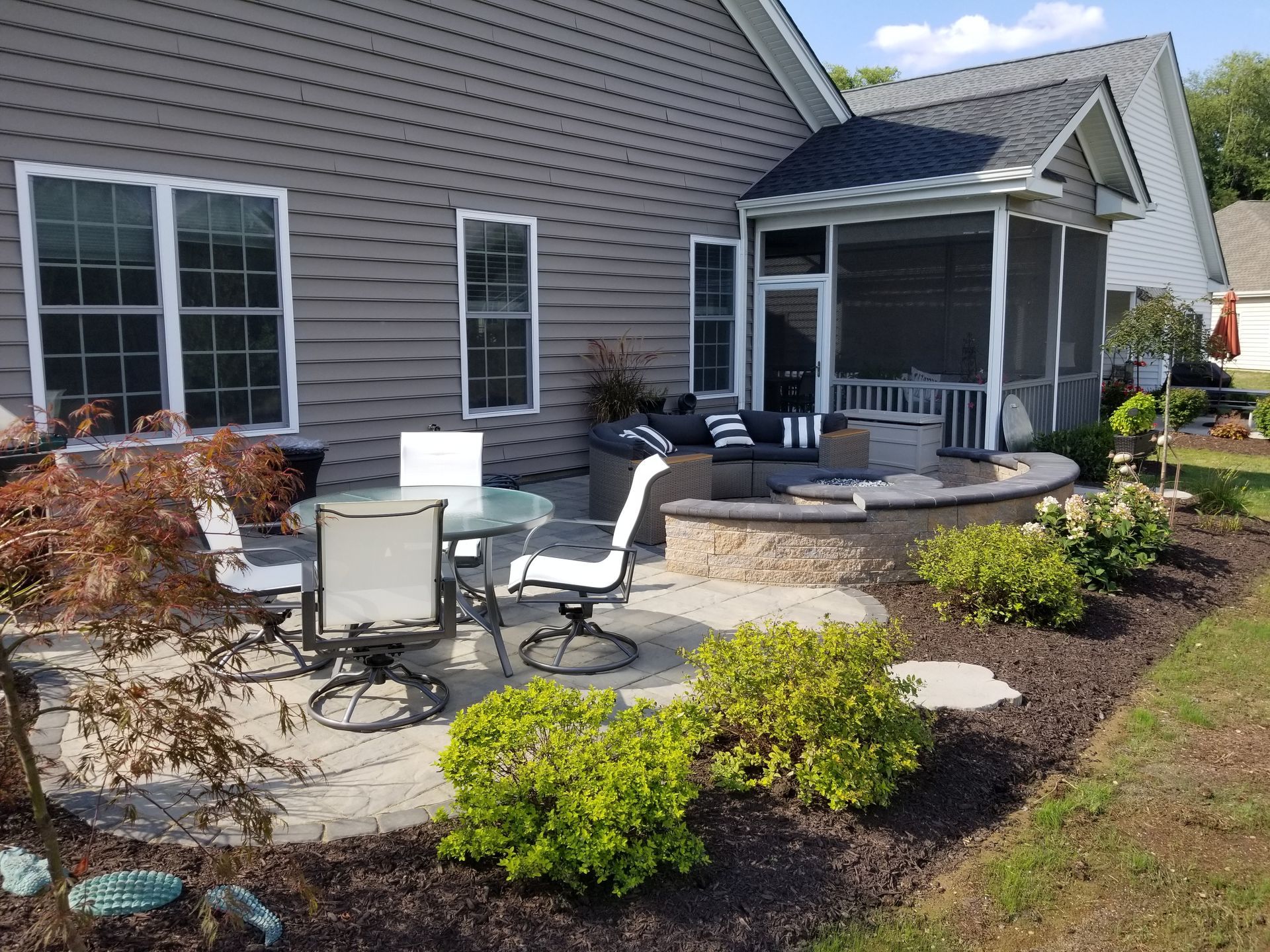 Back patio seating area with chairs, table, and potted plants beside a gray house