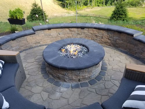 Outdoor stone fire pit with circular seating area and black cushioned chairs overlooking a grassy yard