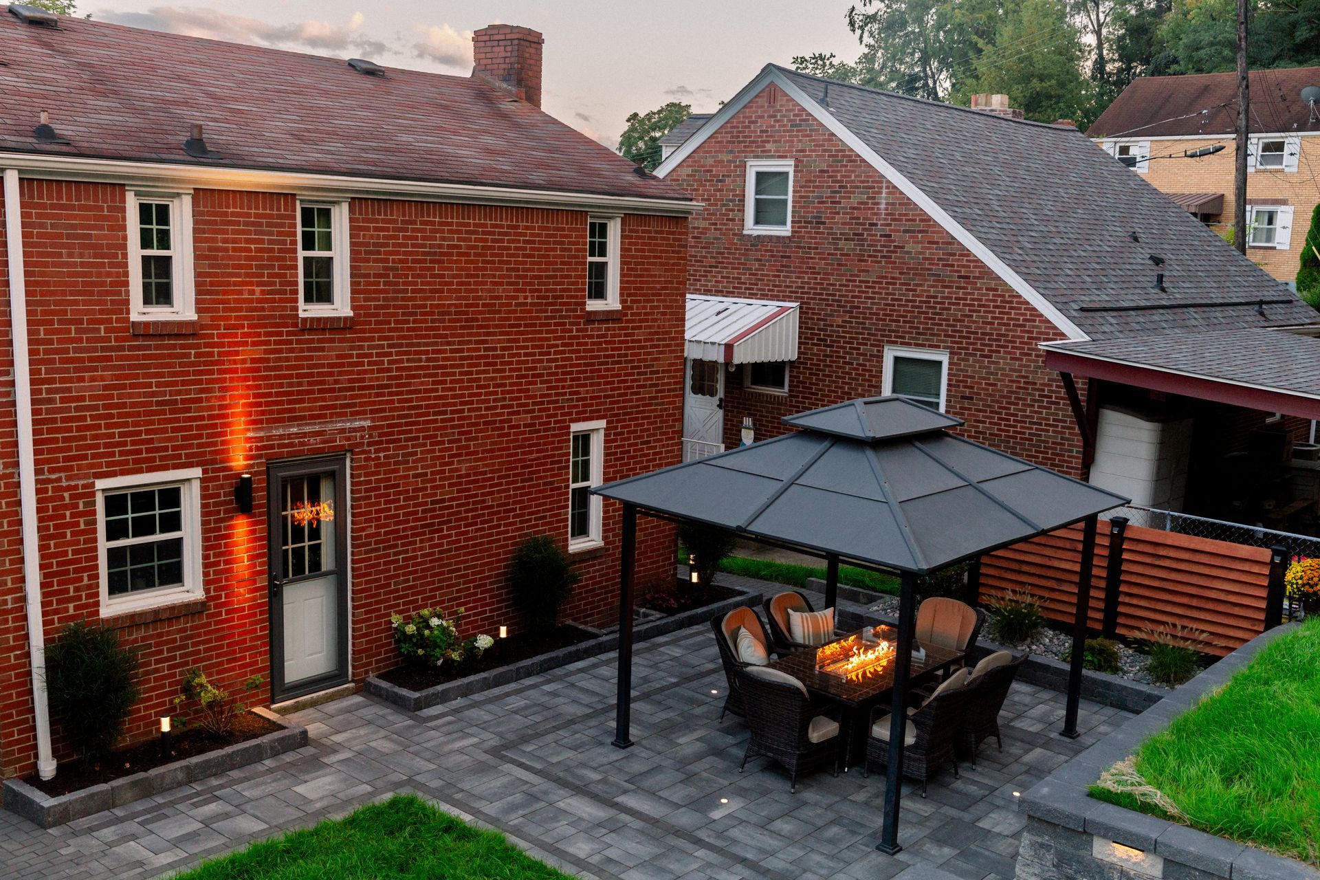 Backyard patio with umbrella, seating, and lit brick houses at dusk