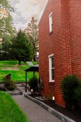 Brick house beside a paved path, with green lawn and trees in a quiet garden setting.