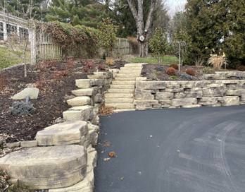Stone retaining wall and steps beside a curved paved driveway in a landscaped yard.