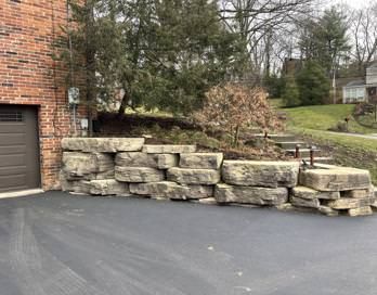 Stone retaining wall blocks stacked beside a driveway next to a brick house and wooded yard.