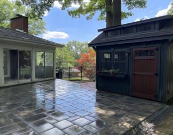 Rainy patio with wet stone pavers between a glass sunroom and a dark shed under trees