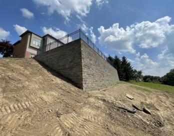 Steep dirt slope beside a stone retaining wall and house under a blue sky with clouds