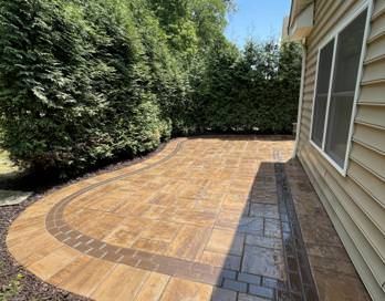 Paver patio beside a house, bordered by dense green hedges and a paver walkway strip.
