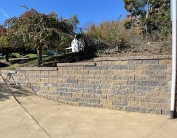 Stone retaining wall beside a sidewalk, with trees and shrubs on a sunny hillside