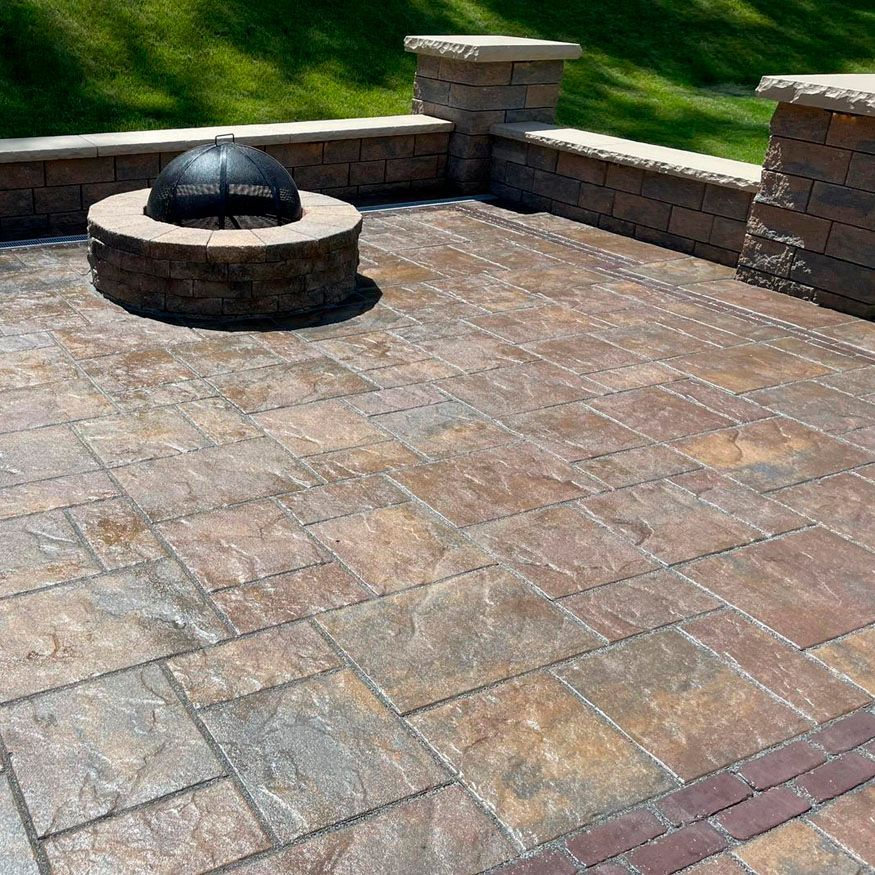 Stone patio with a round fire pit and low brick seating wall in a sunny backyard