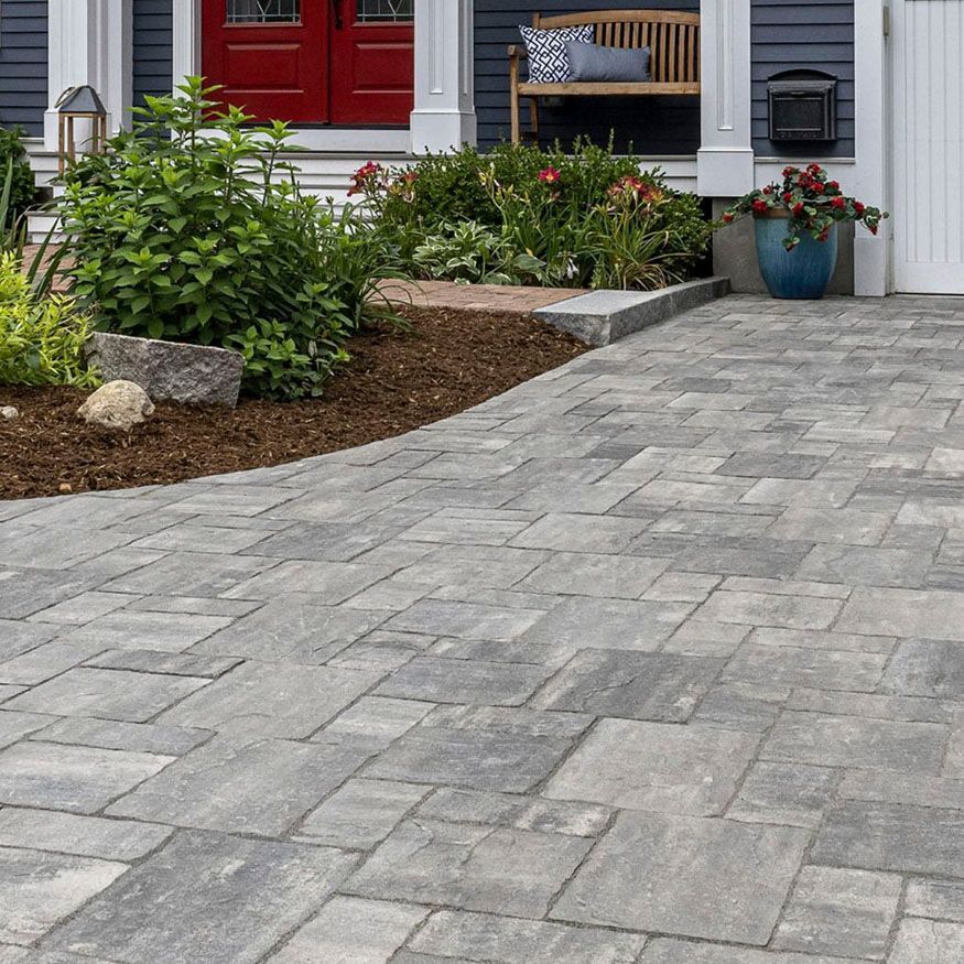 Gray paver walkway leading to a house entrance with flower beds, shrubs, and a bench on the porch