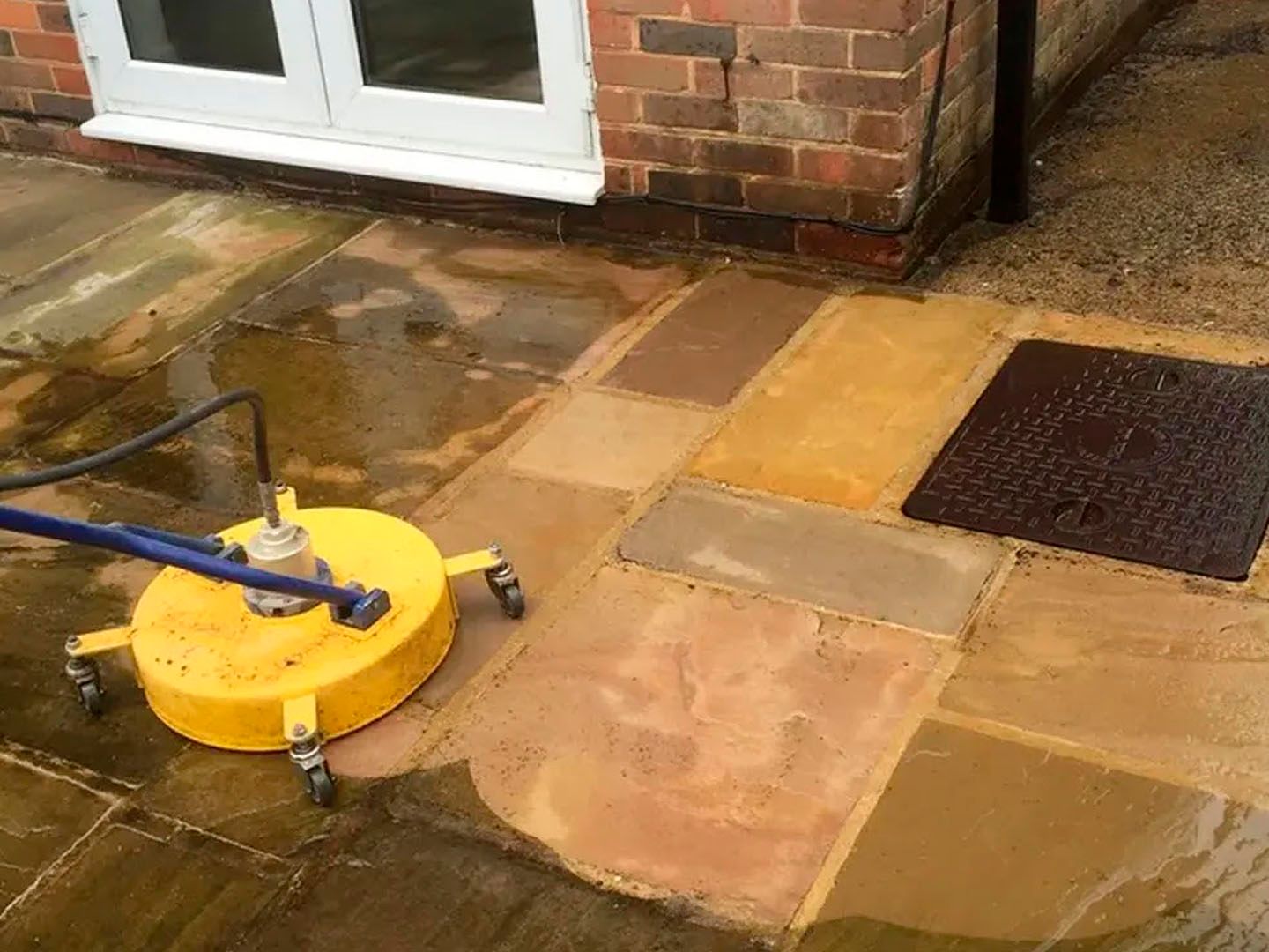Yellow pressure washer on a wet stone patio beside a brick house and drain cover
