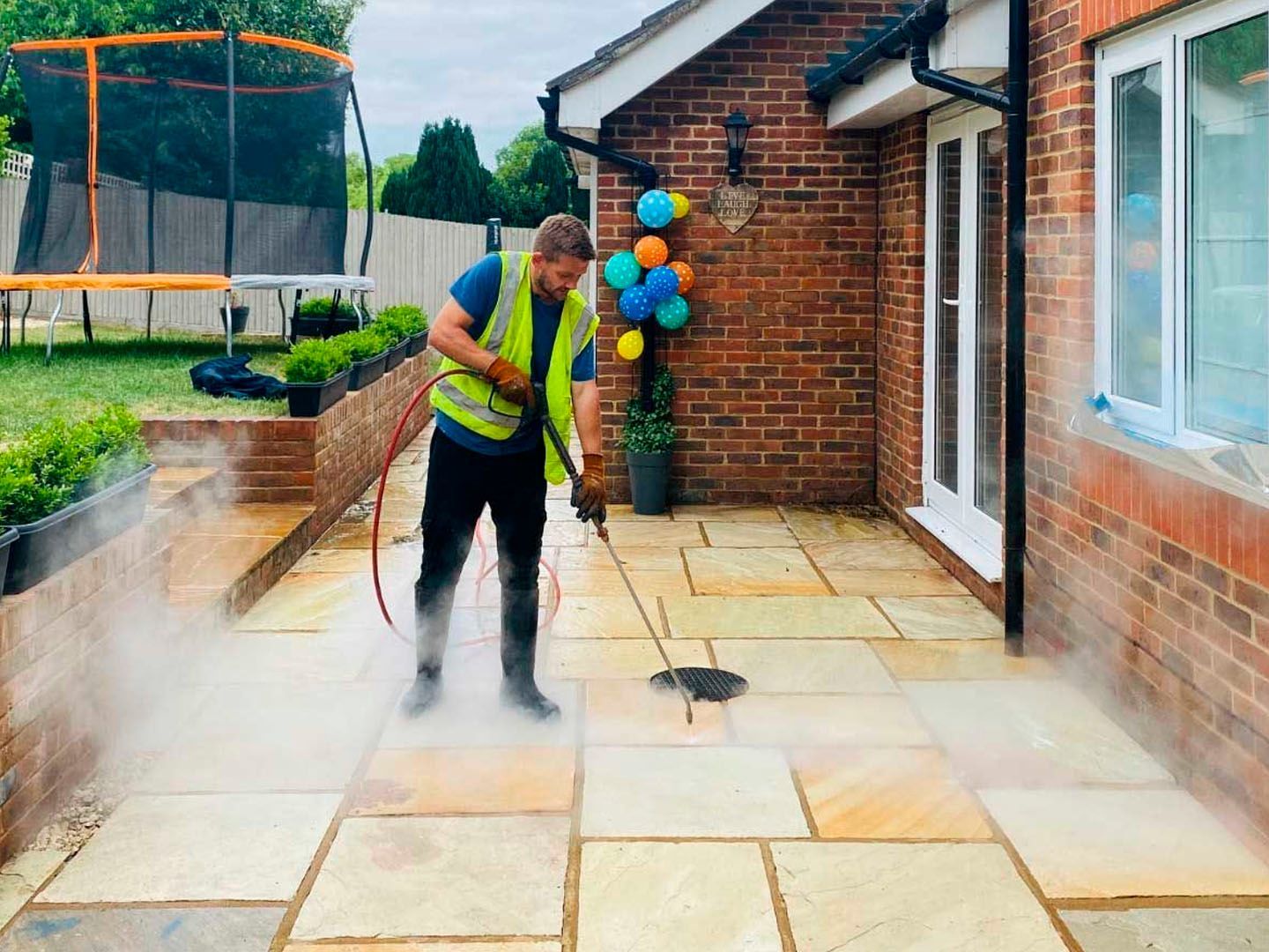Person pressure-washing a patio beside a brick house, with balloons by the door.