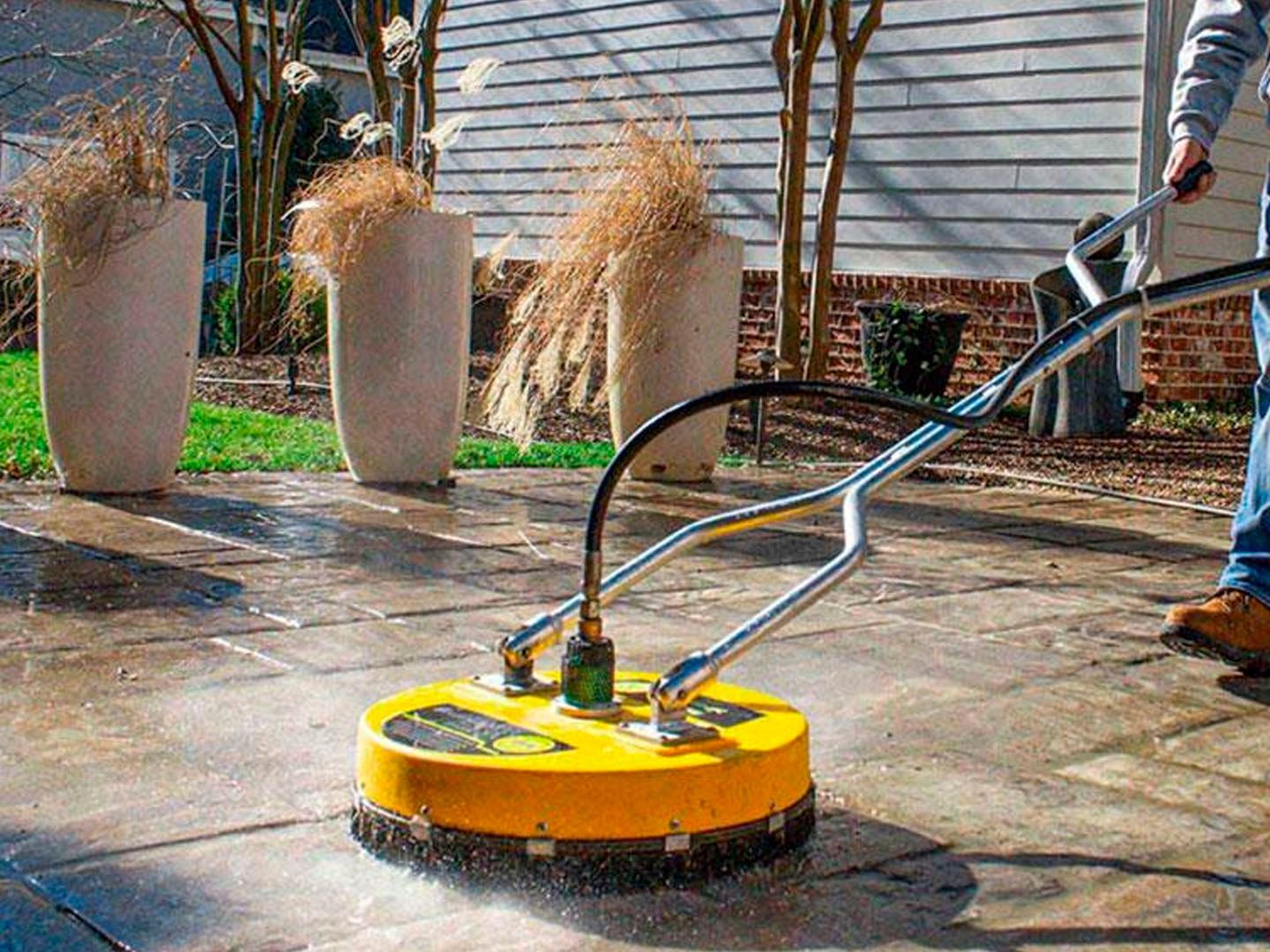 Yellow pressure washer cleaning a concrete patio near tall planters and a house exterior
