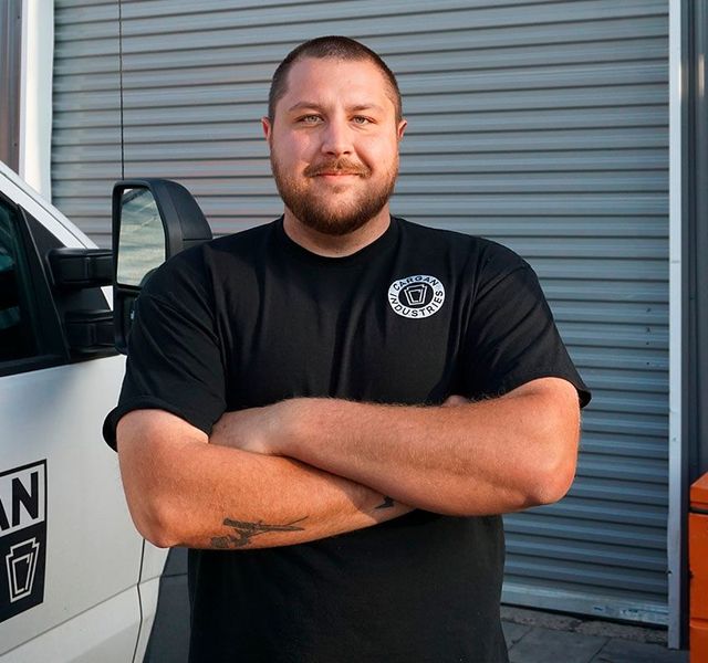 Man standing with arms crossed beside a vehicle, in front of a gray roll-up door.