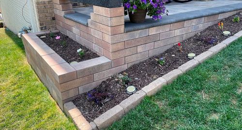 Brick garden border with mulch bed, small plants, and green lawn beside a porch steps.