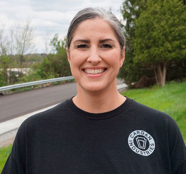 Smiling person in a black T-shirt standing outdoors beside a road and trees