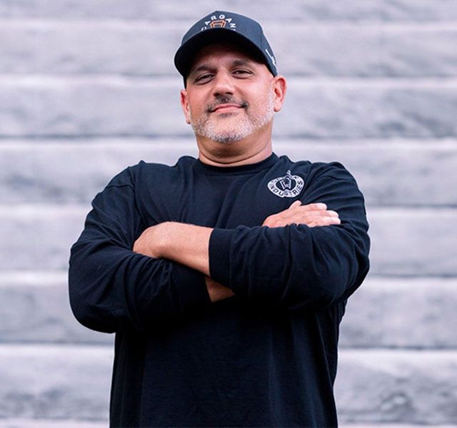 Man standing with arms crossed in a black shirt and cap against a gray wall