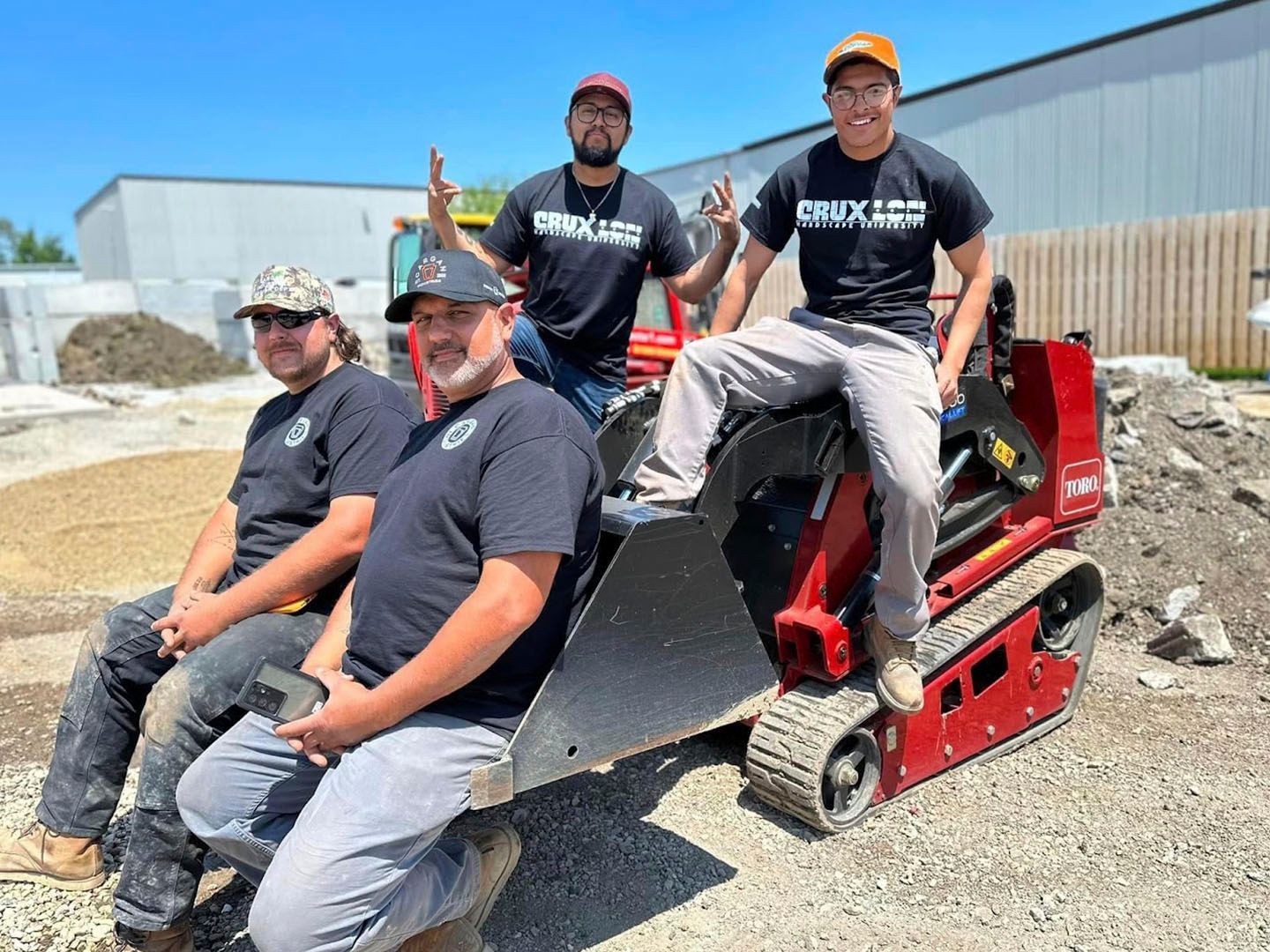 Four workers posing on a red skid-steer loader in a gravel lot near a warehouse
