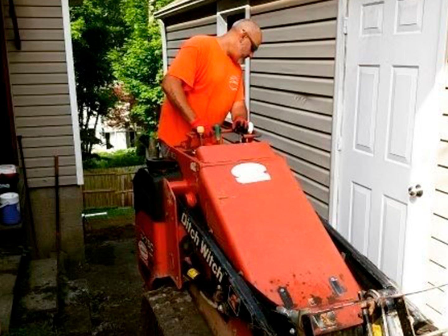 Man in orange shirt operating a red lawn machine in a narrow side yard between houses