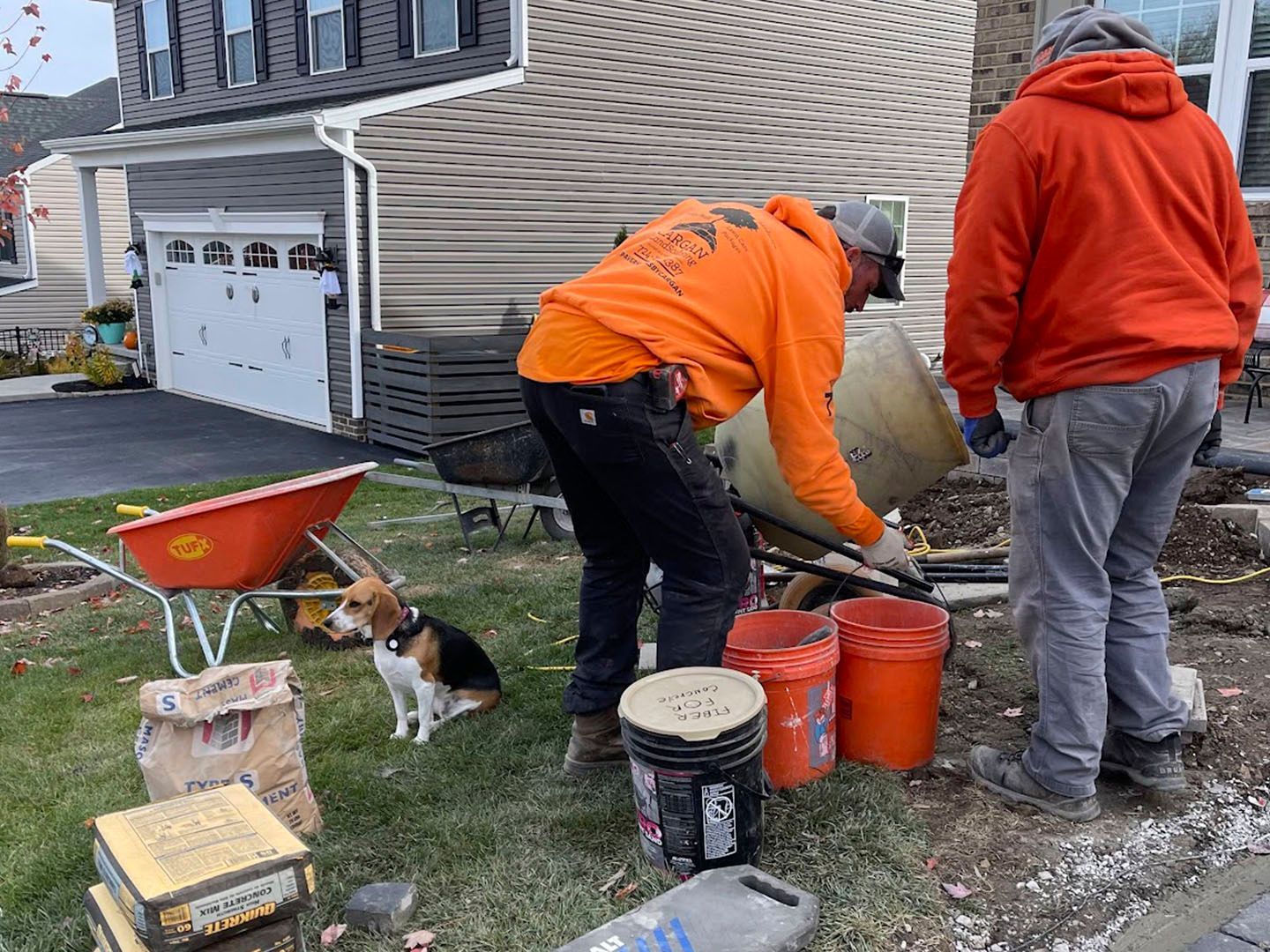 Two workers in orange jackets mixing concrete beside buckets and a wheelbarrow in a driveway, with a dog nearby