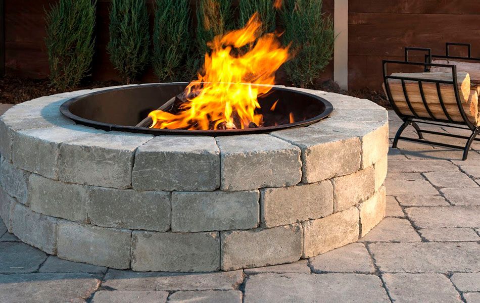 Stone fire pit with bright flames on a patio, with outdoor chairs nearby