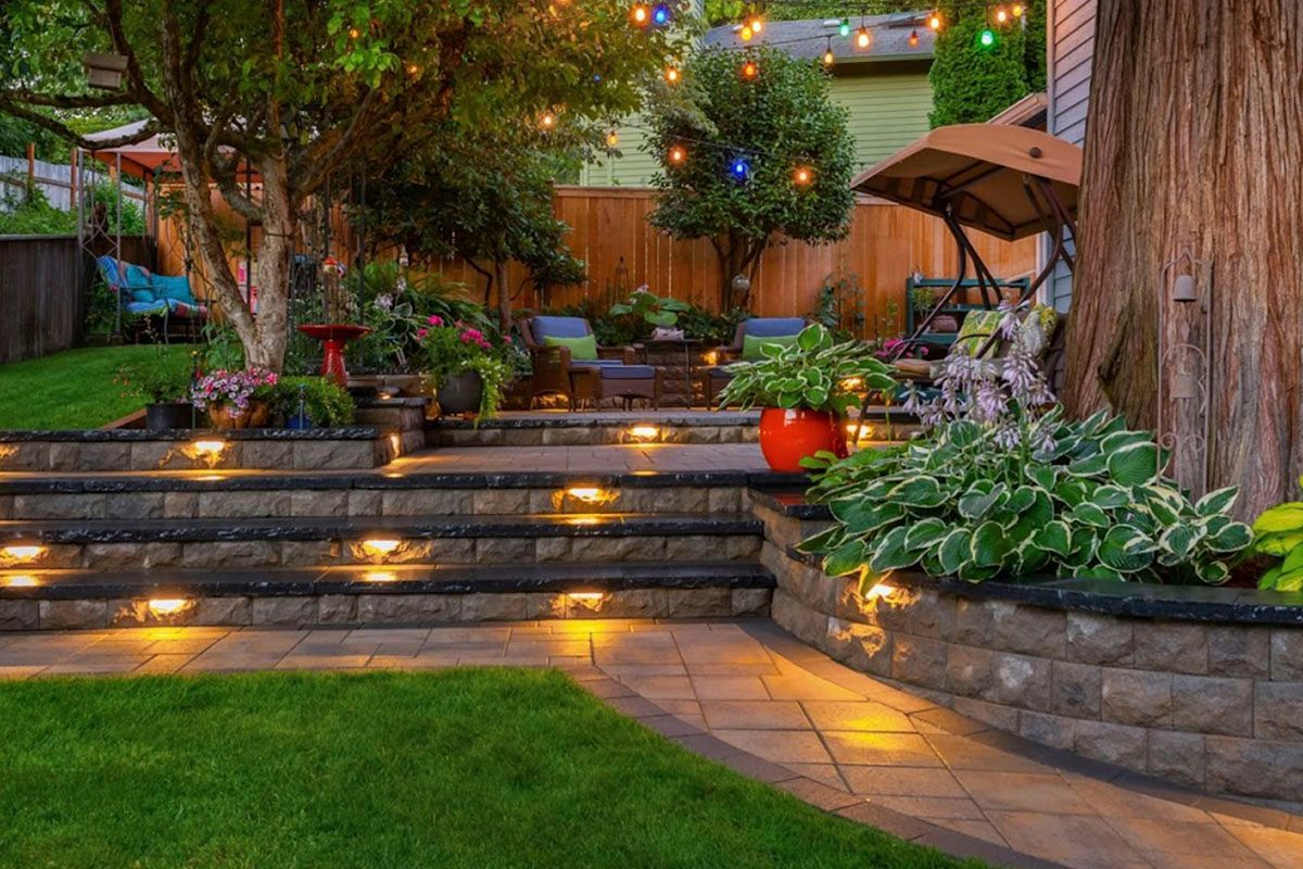 Illuminated garden patio with stone steps, potted plants, and string lights at dusk