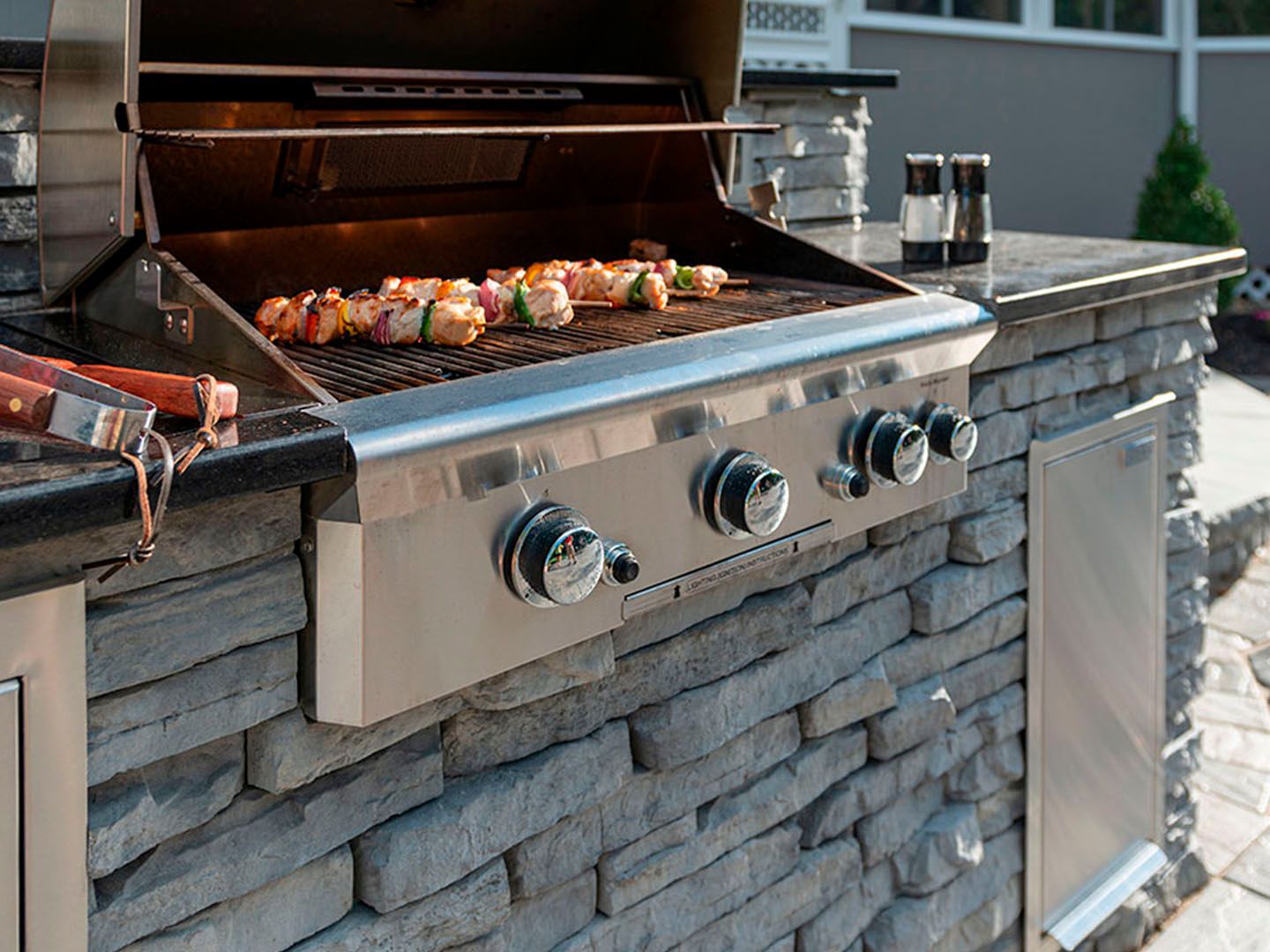 Built-in outdoor gas grill with skewers cooking on the grates beside a stone counter