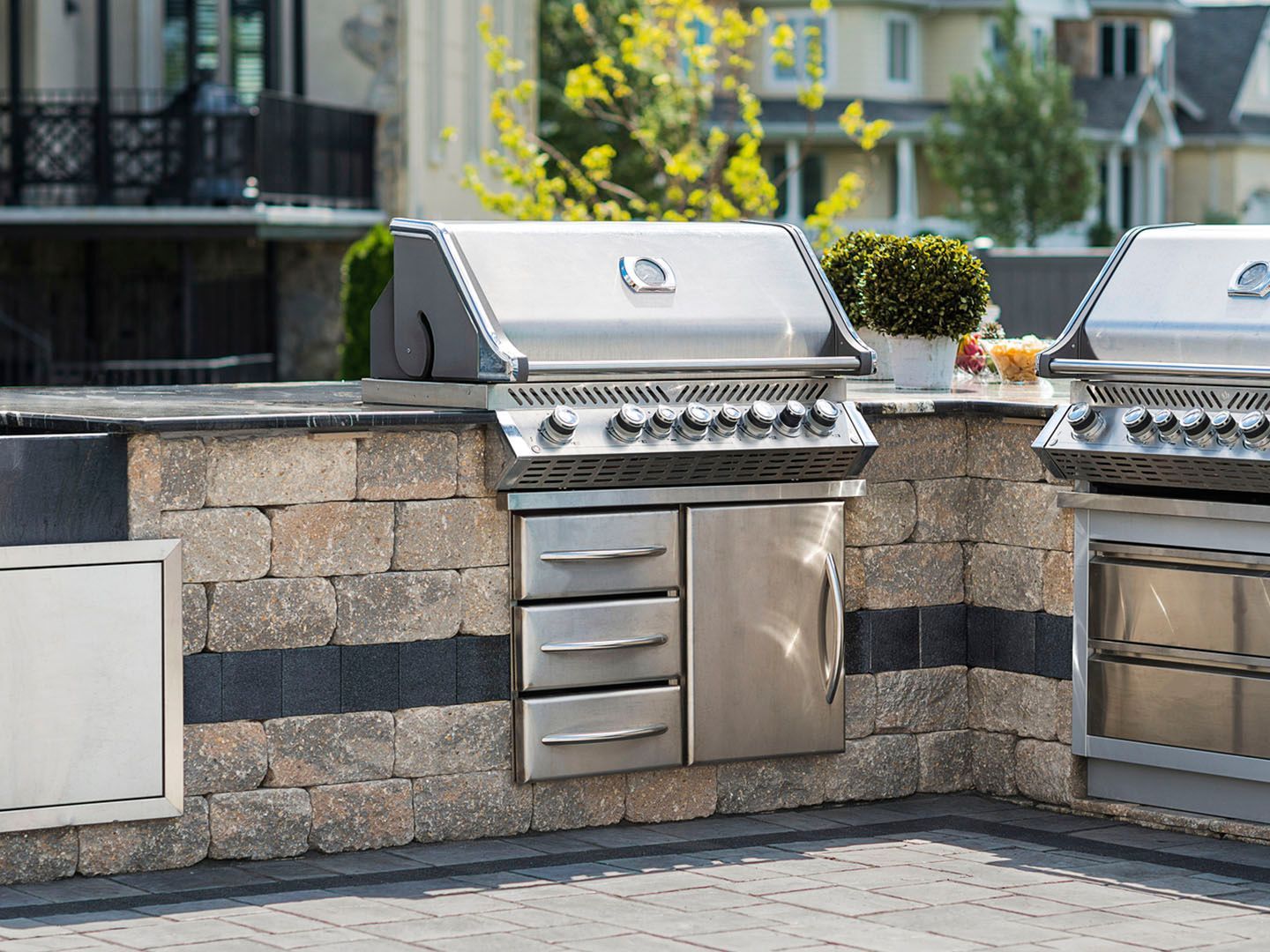 Outdoor stainless-steel gas grills built into a stone wall on a patio with apartment buildings in the background