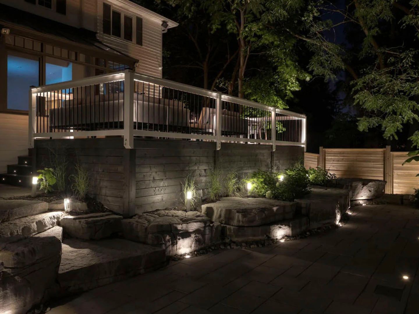 Nighttime backyard deck with white railing, stone retaining wall, and soft landscape lighting