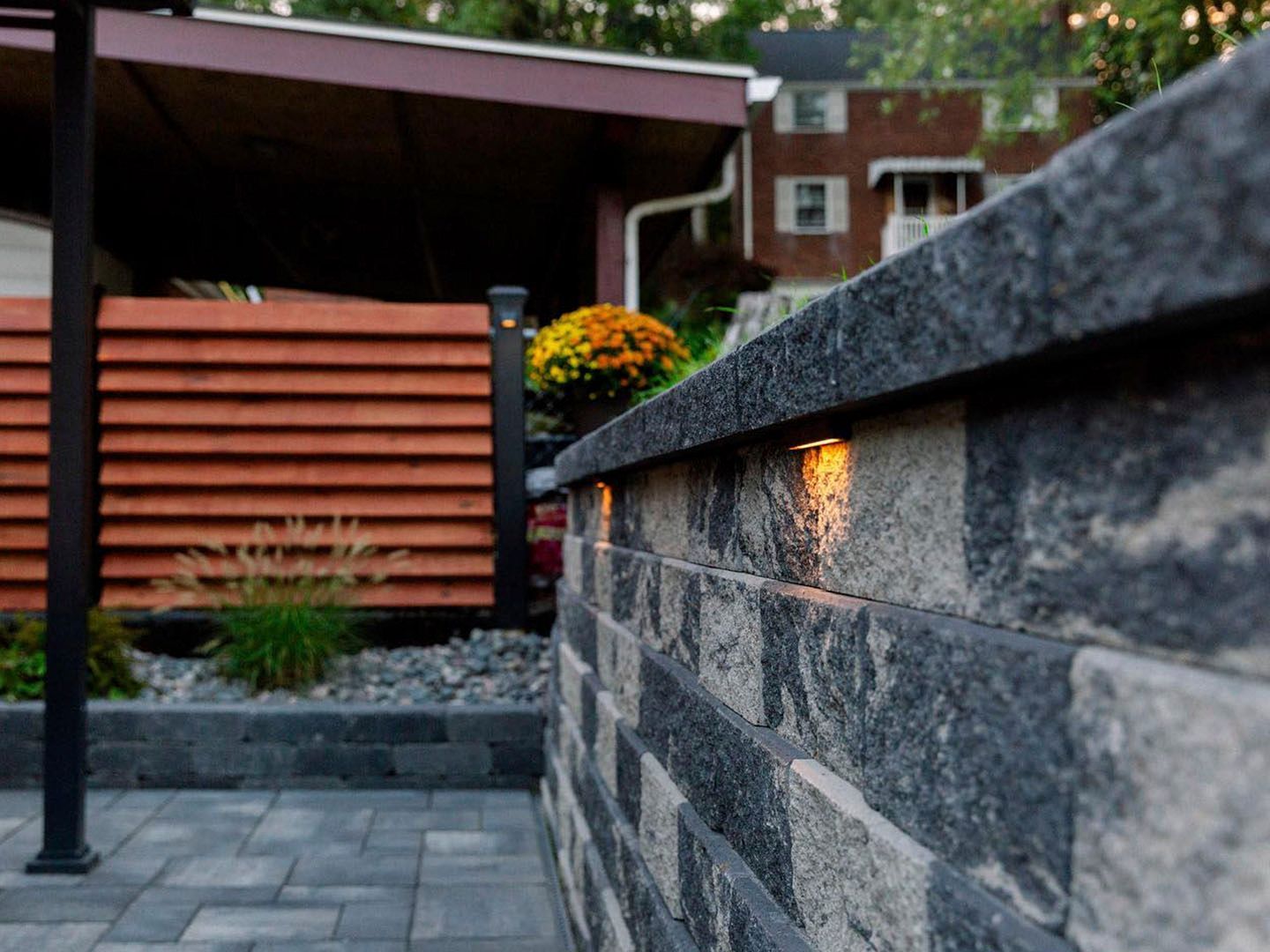 Stone wall with a lit sconce beside a paved courtyard and wooden fence