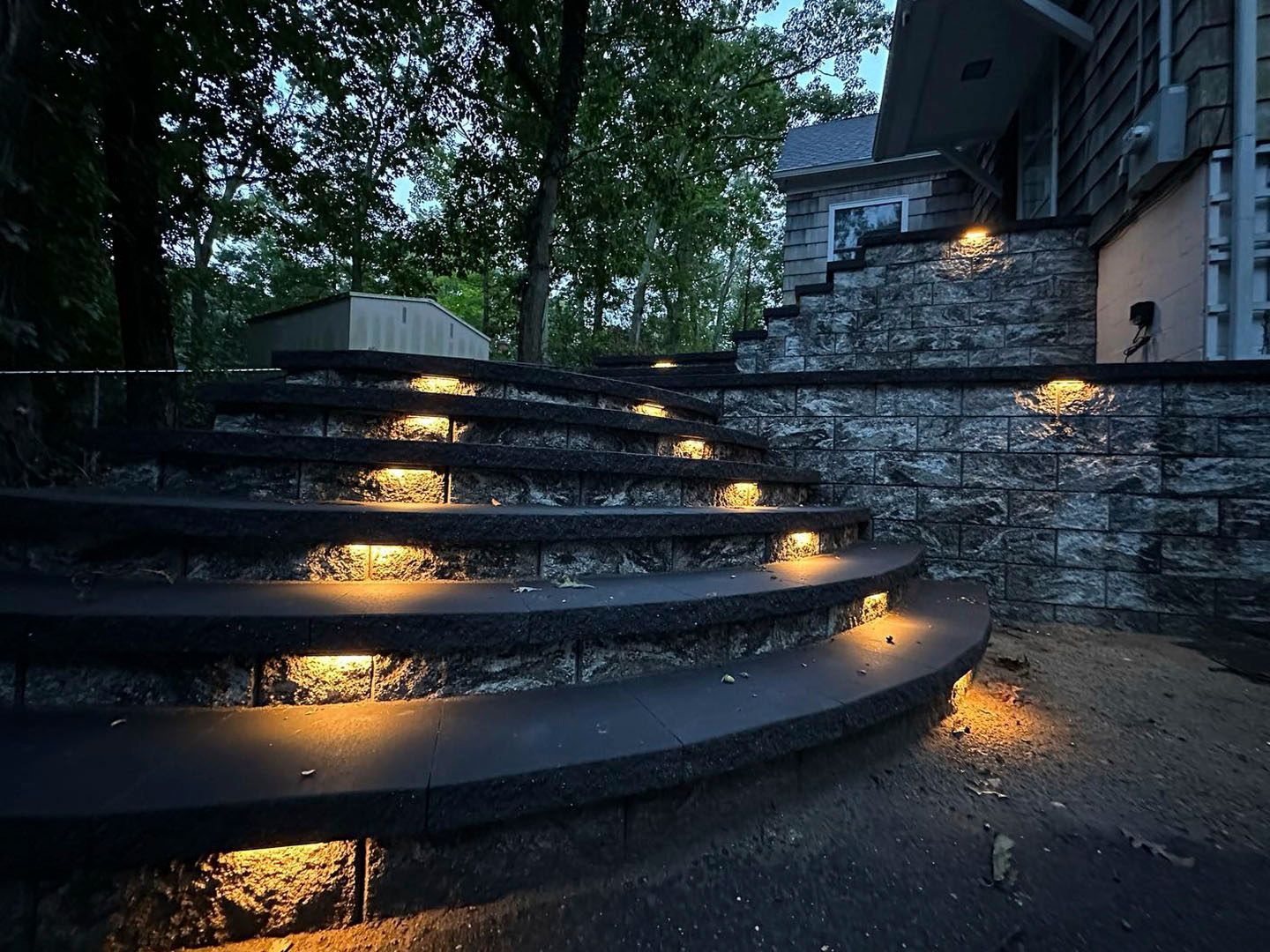 Curved stone steps with glowing lights at dusk beside a house and trees