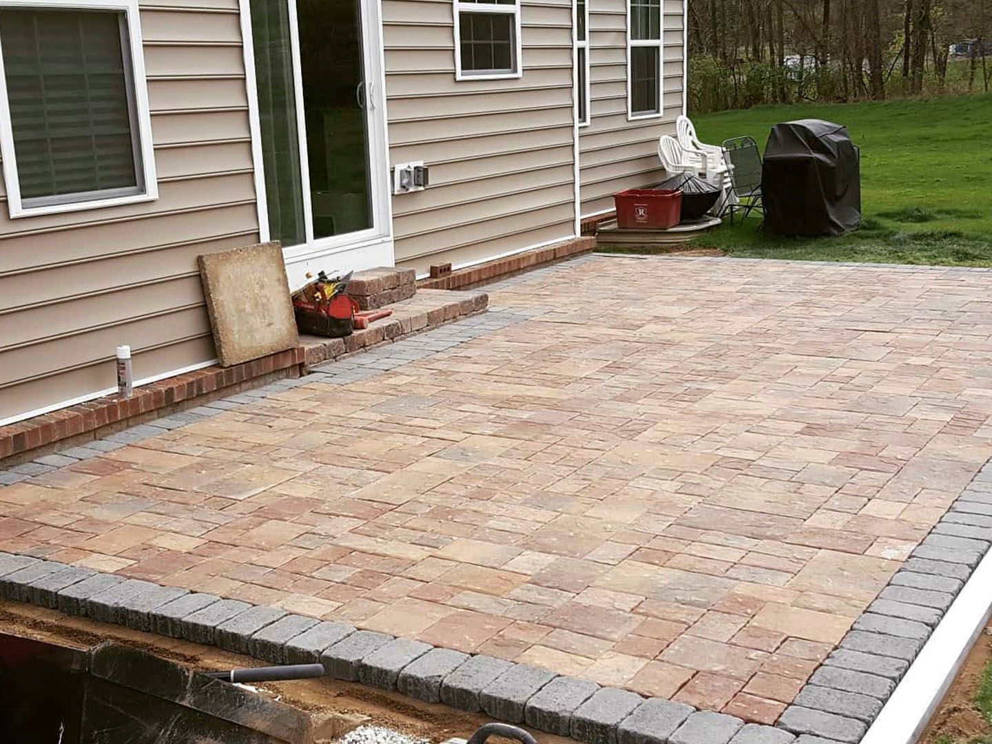 Brick patio beside a house, bordered by gray pavers, with a grassy yard and outdoor equipment nearby
