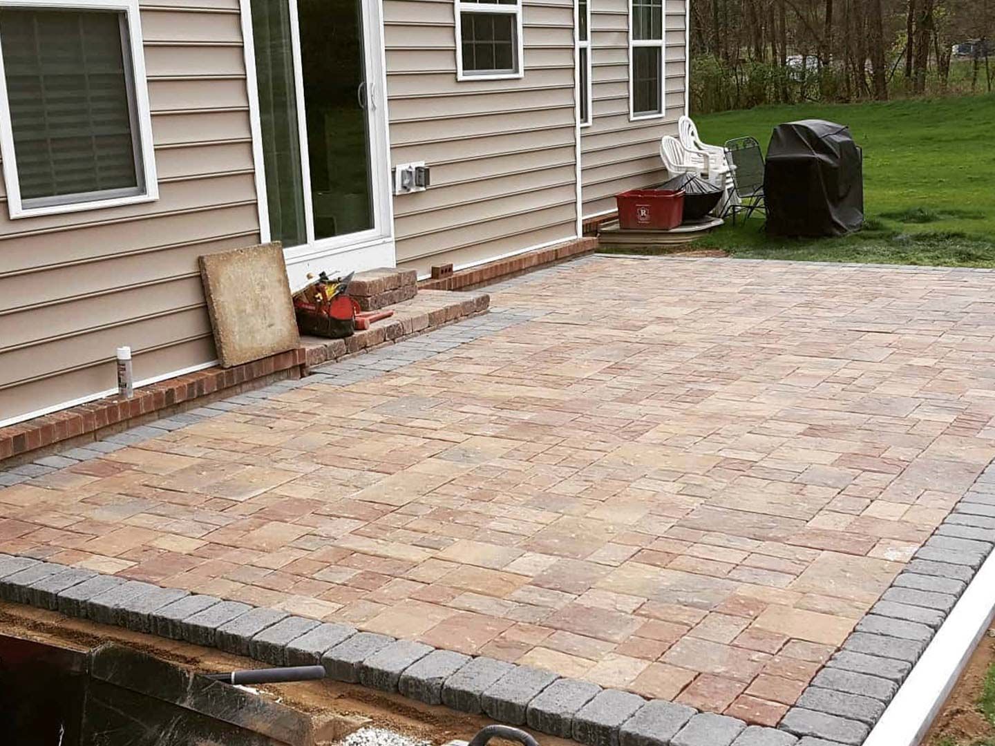 Backyard brick patio beside a house with beige siding and a grill on the grass end