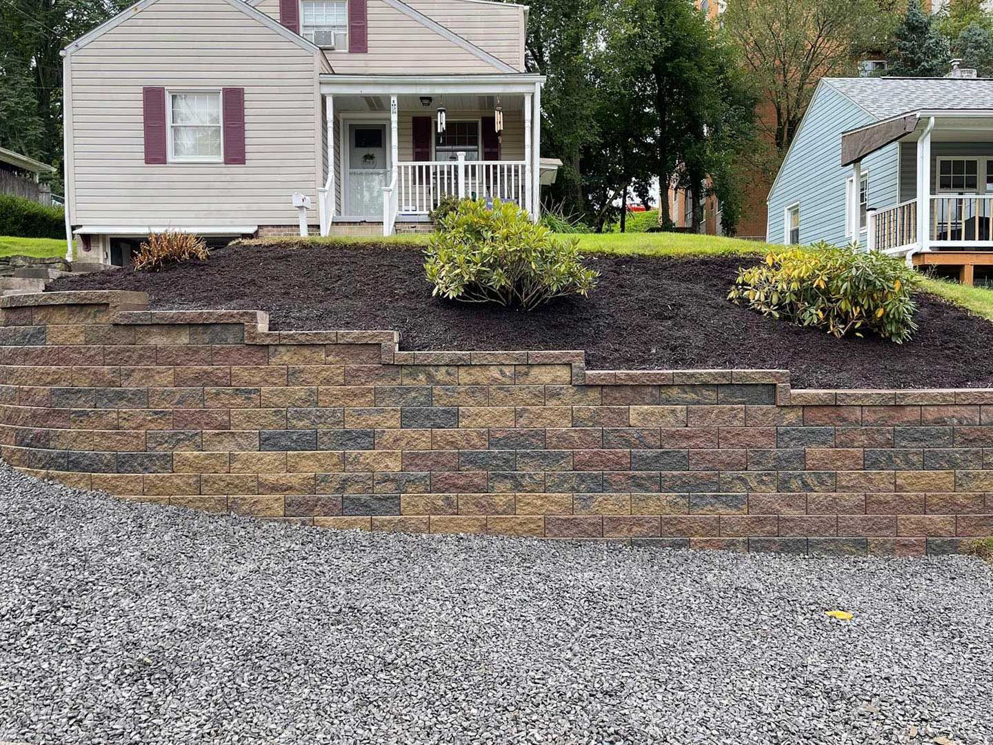Two-story house with a landscaped front yard and stacked stone retaining wall beside a gravel driveway