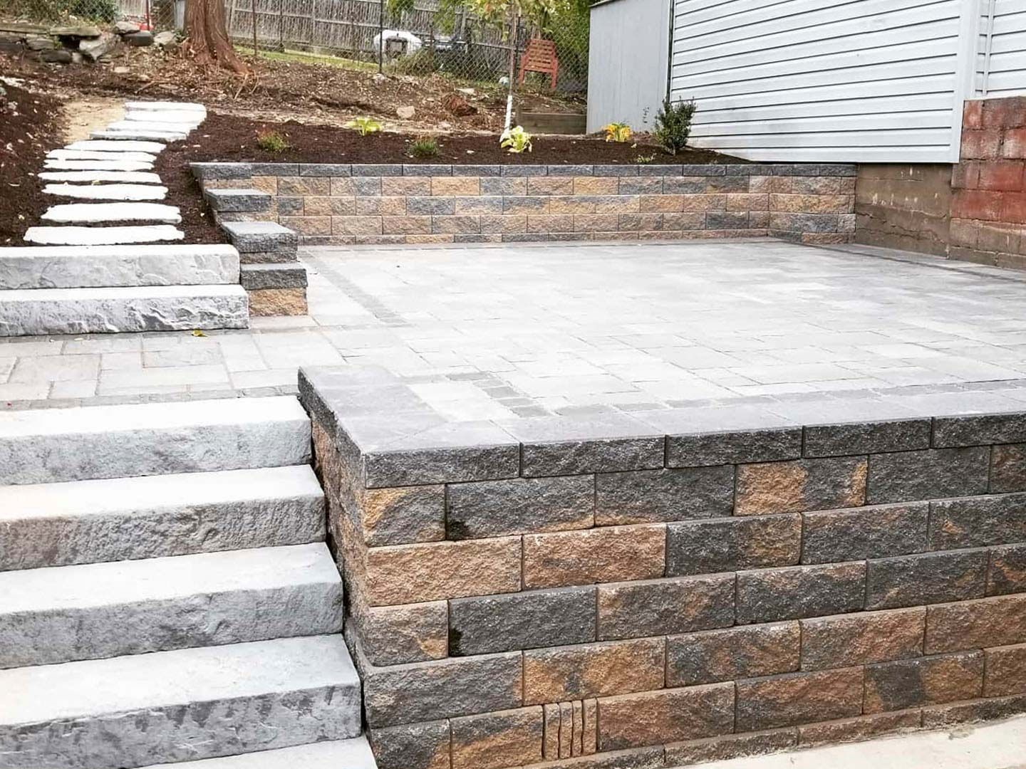 Stone steps and a large paved patio beside a brick retaining wall and white house exterior