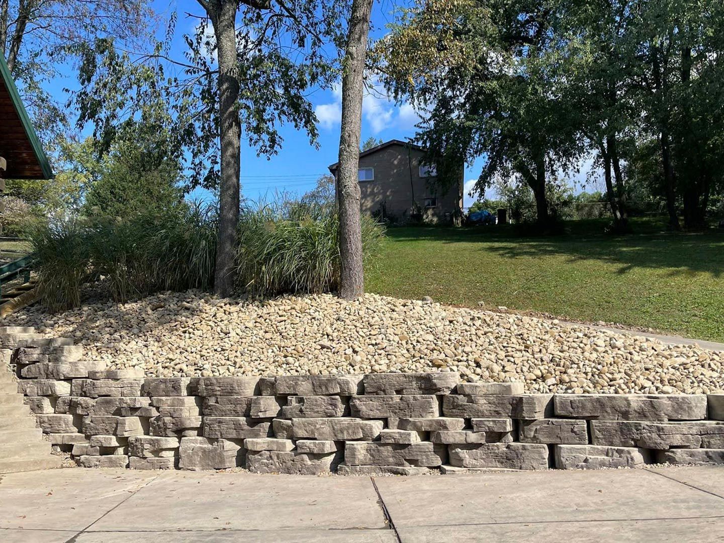 Stone retaining wall below a sloped yard with trees and a house in the background
