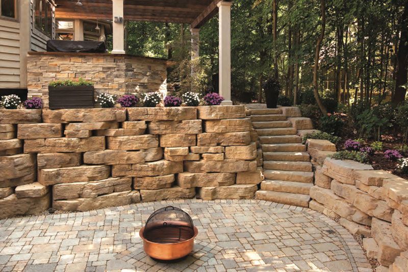 Stone patio with fire pit in a wooded backyard, featuring tiered retaining walls and steps.