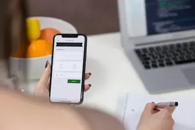 Person holding a smartphone with a login screen beside a laptop on a desk