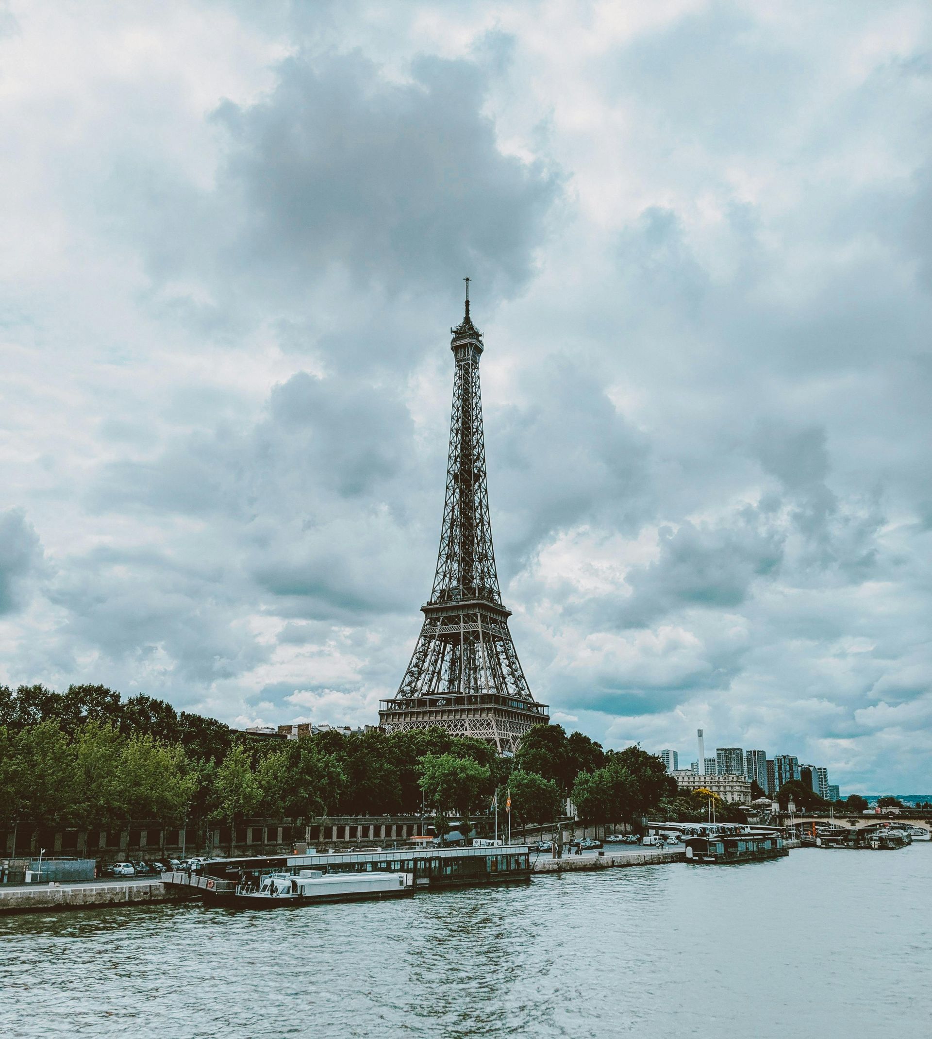 Eiffel Tower rising above the Seine under cloudy skies, with riverbanks and trees in the foreground