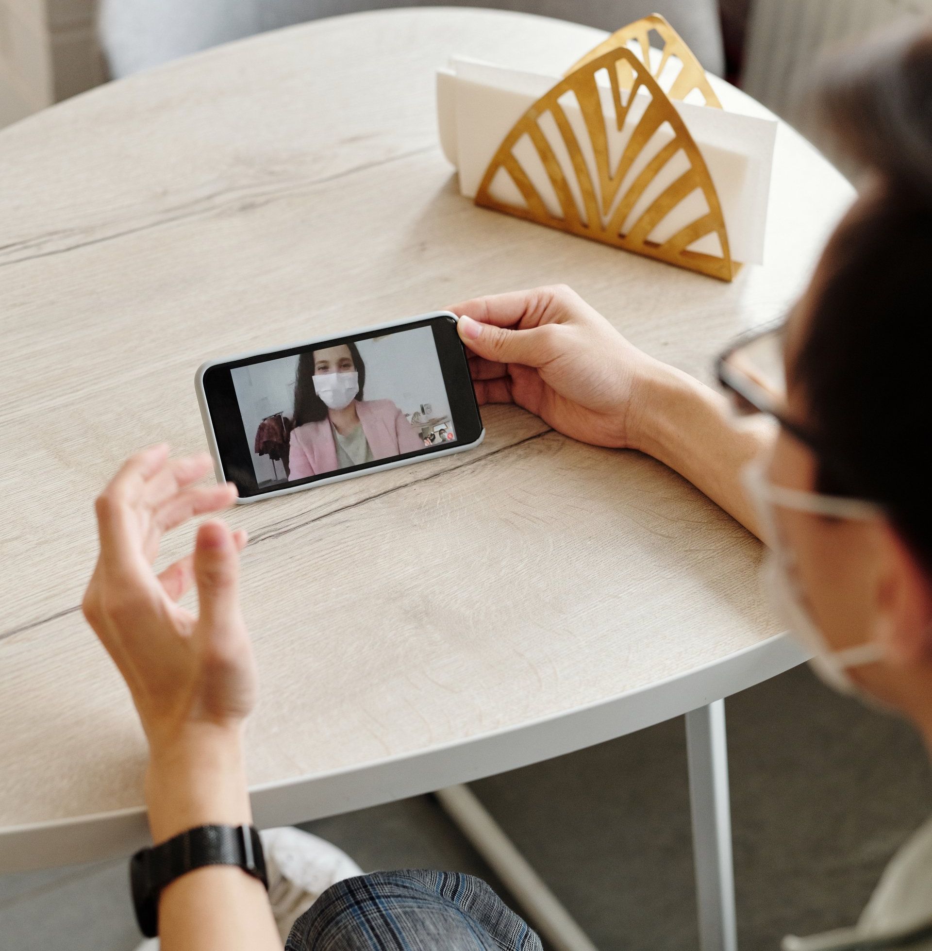 Person video chats on a smartphone across a round table, wearing a mask in a bright room.