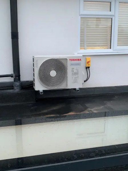 Toshiba air conditioning unit mounted on a white wall, near a window. Black roof below.