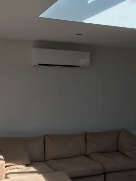 White wall-mounted air conditioner above a beige sectional sofa in a room with a skylight.