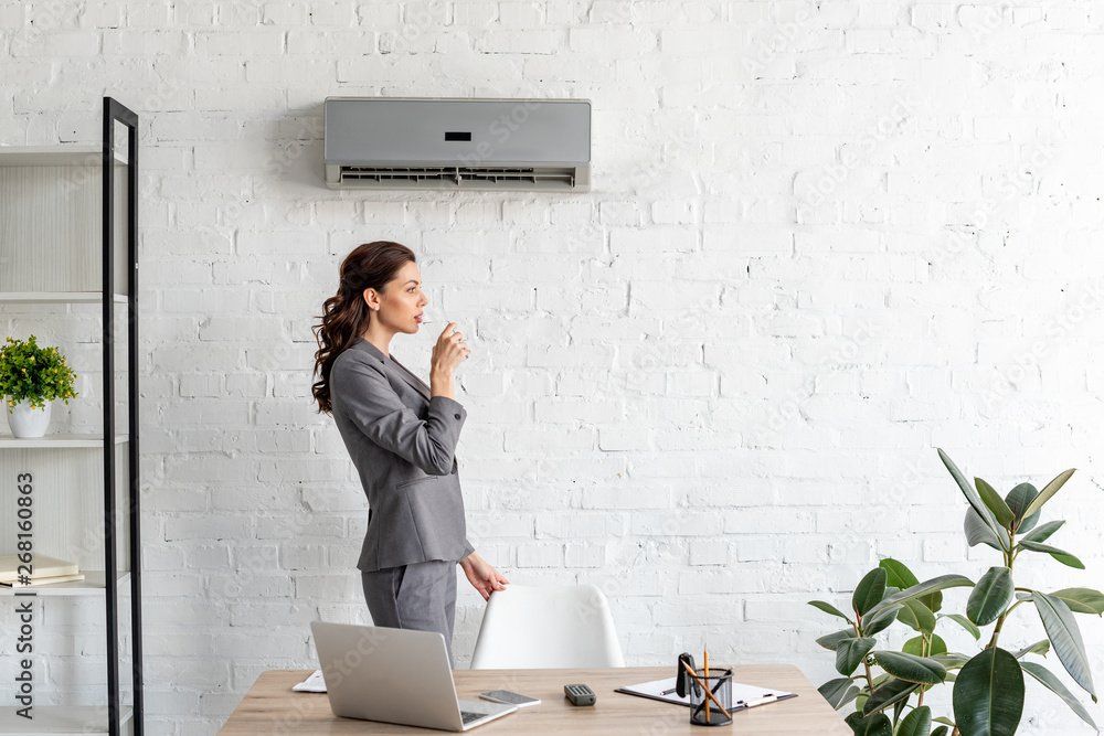 Woman in a gray suit stands in an office, holding a glass of water under an air conditioner.