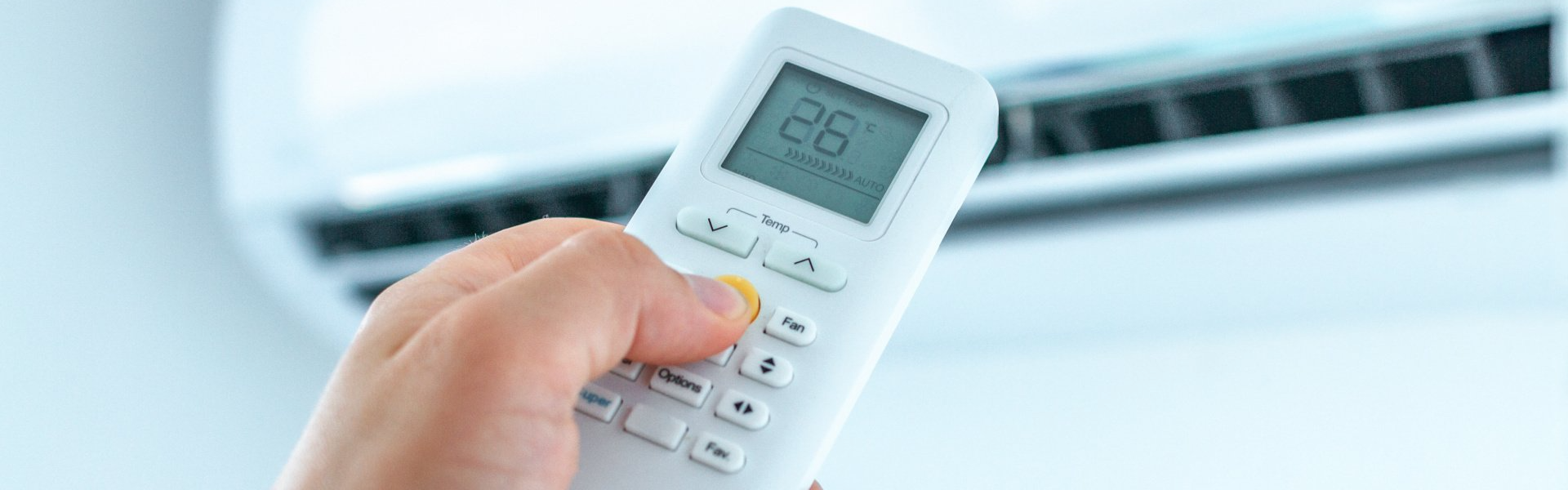 Hand holding a white remote, pointing towards an air conditioner. The display shows the number 26.
