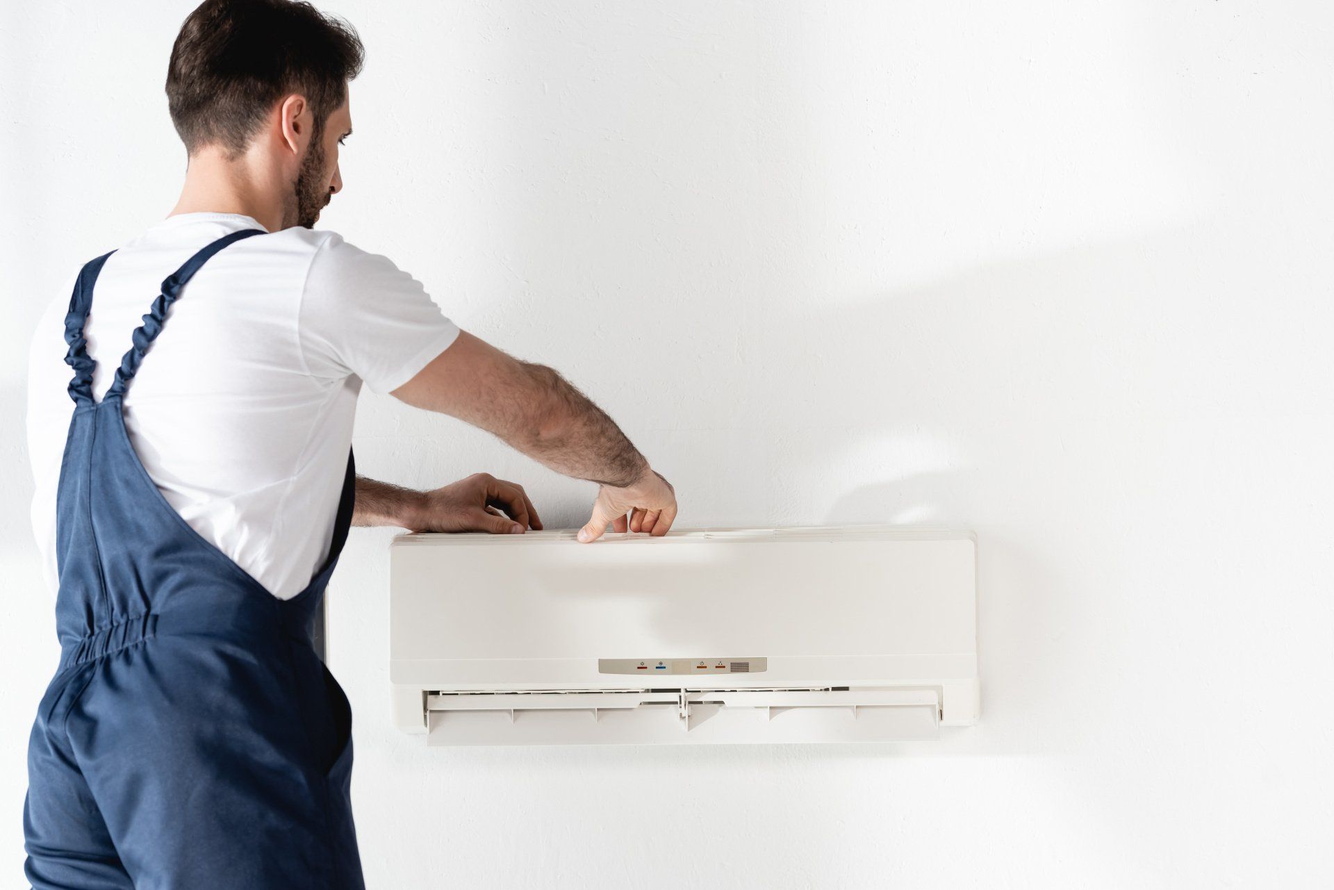 Man in blue overalls servicing a white wall-mounted air conditioner.