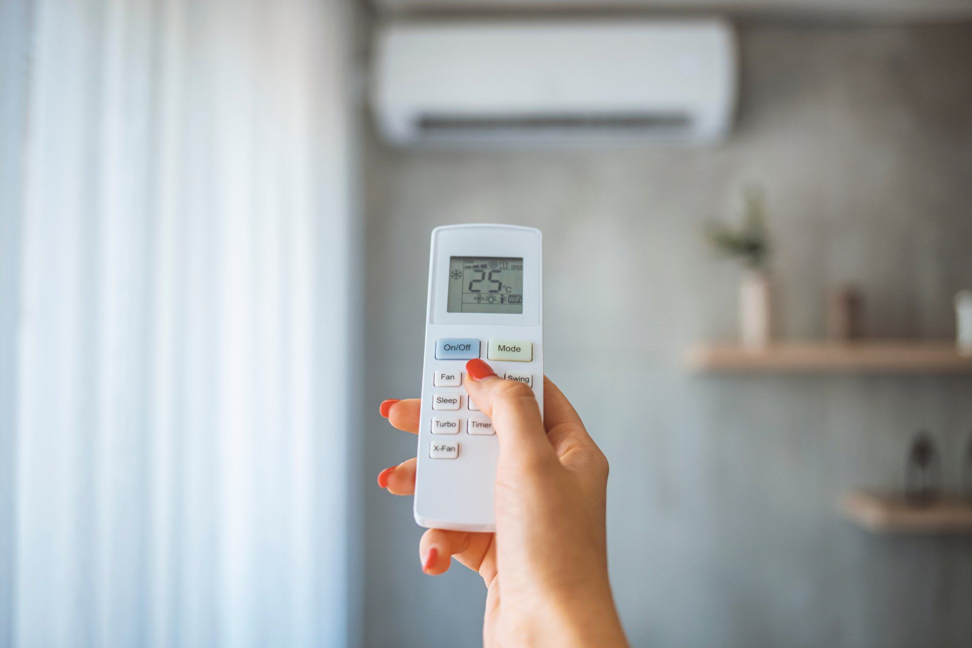 Hand holding air conditioner remote, adjusting settings, unit mounted on wall.