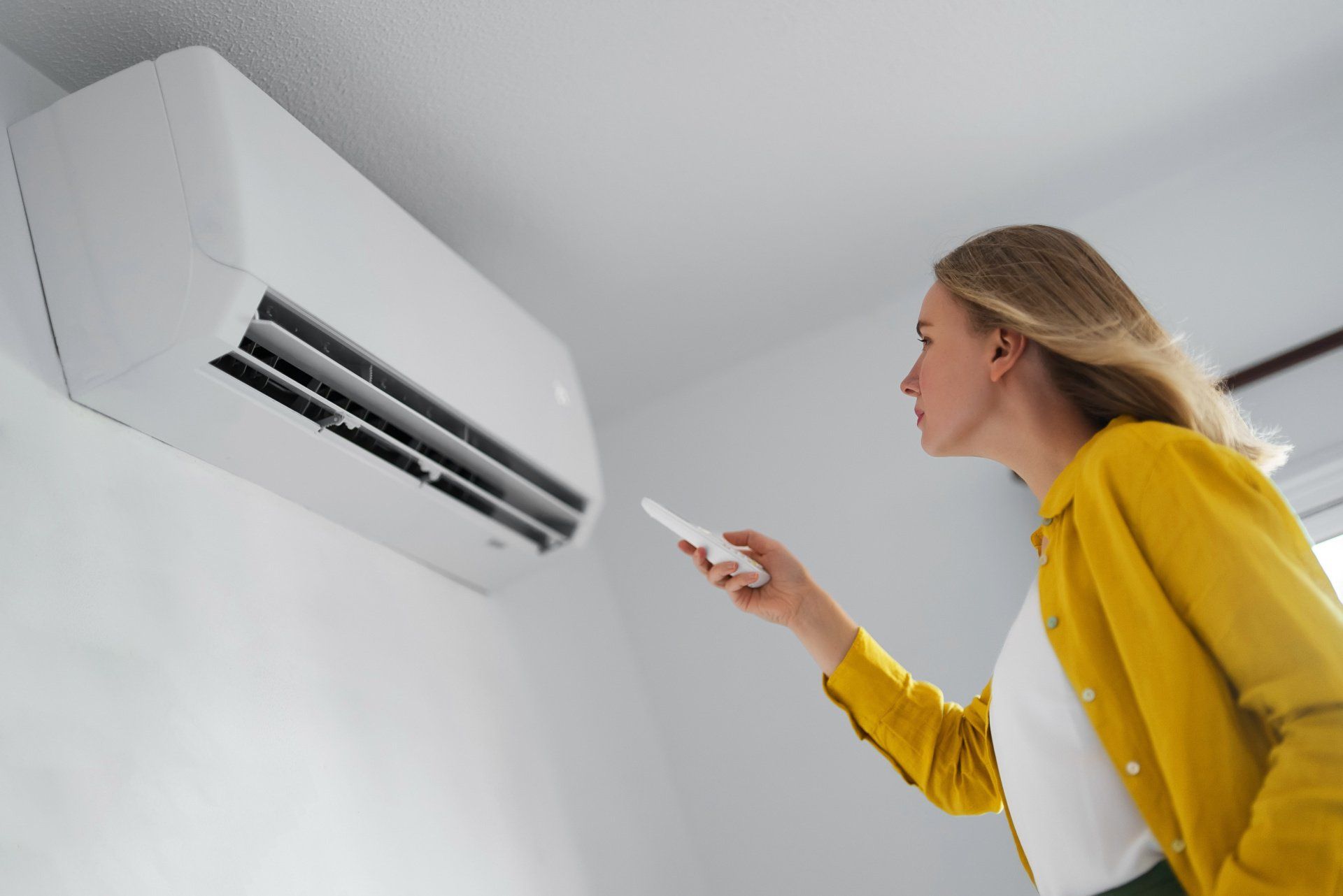 Woman in yellow jacket uses remote to operate an air conditioner mounted on a white wall.