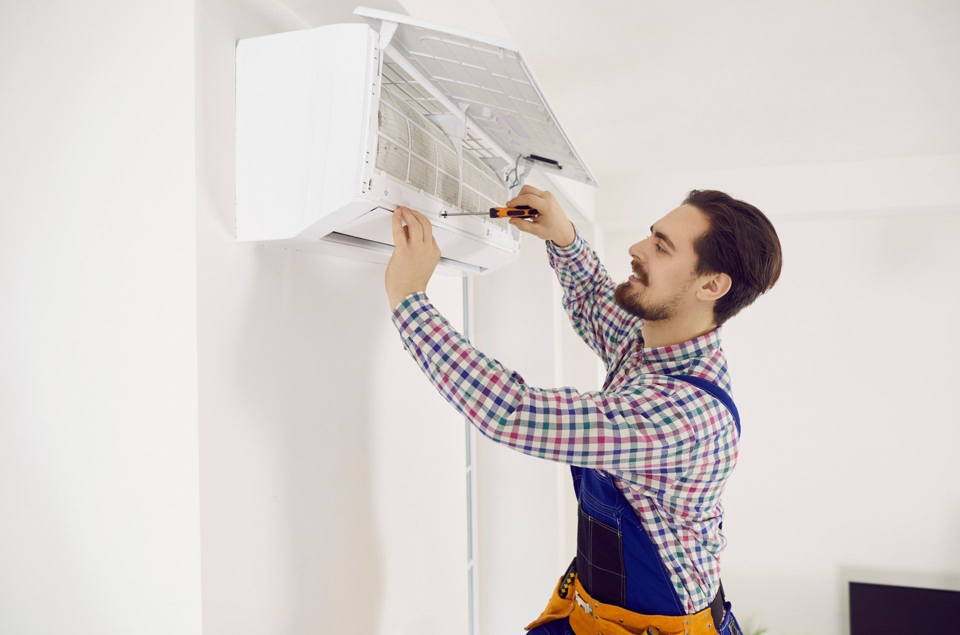 Man in plaid shirt and overalls, fixing a wall-mounted AC unit with a screwdriver. White room setting.