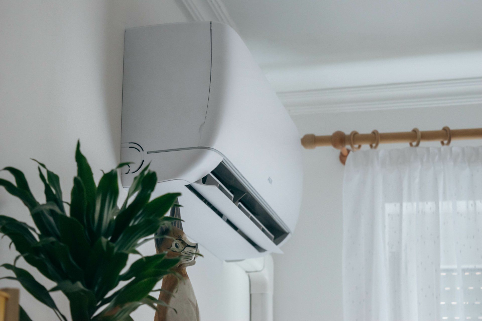 White air conditioning unit mounted on a white wall above a plant, near a window with curtains.