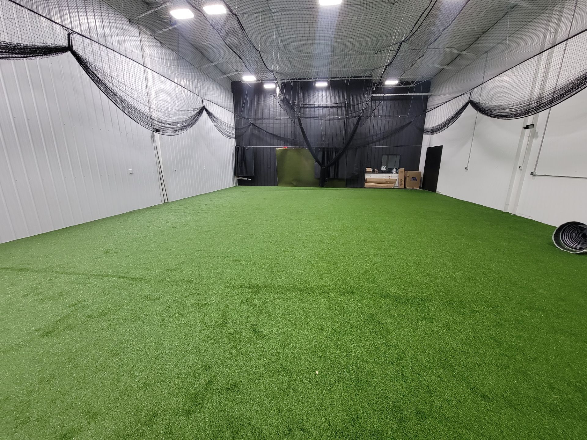 Indoor sports training facility with green turf, netted walls, and a pitching mound at the far end